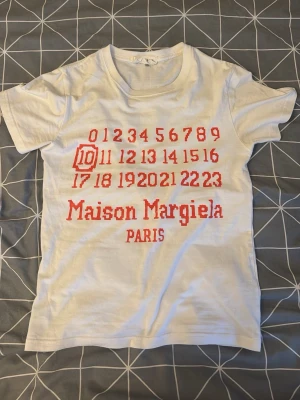 Maison Margiela vit t-shirt med tryck - Maison Margiela t-shirt i vitt med ikoniskt rött tryck framtill och logga. Klassisk passform med korta ärmar och rund hals. Tillverkad i mjuk bomull som känns skön mot huden. Perfekt för dig som gillar streetstyle och designerplagg.