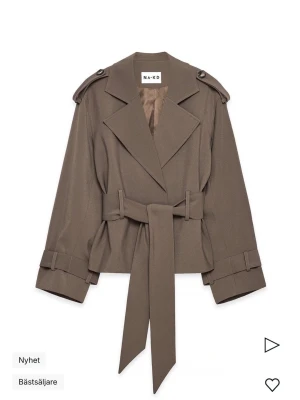 Trenchcoat från NA-KD - Stilren brun trenchcoat från NA-KD med breda slag, axelklaffar och knytskärp i midjan. Jackan har långa ärmar med spänndetaljer vid ärmslut och en rak, något kortare passform. Perfekt för dig som gillar trendiga och klassiska plagg.💓 Nypris: 1099