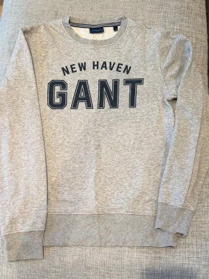 Grå sweatshirt från Gant - Snygg grå sweatshirt från Gant med trycket 'NEW HAVEN GANT' på bröstet. Klassisk rund halsringning och ribbade muddar vid ärmslut och nederkant. Perfekt för en avslappnad och stilren look.