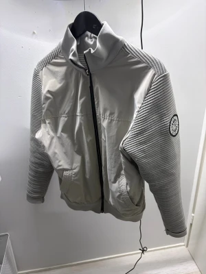 Grå vindjacka med strukturmönster - Snygg grå vindjacka från canada goose med hög krage och hel dragkedja framtill. Jackan har släta paneler framtill och bak samt ärmar och axlar i ribbat strukturmönster. Canada goose-logga på vänster ärm. Perfekt för en stilren och sportig look.