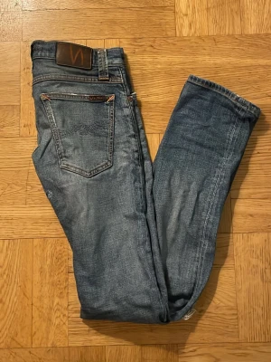  Nudie Jeans - Säljer ett par blå jeans från Nudie Jeans med slitningar och hål på benen för en cool, avslappnad look. Klassisk femficksmodell med smal passform och detaljerade sömmar på bakfickorna. Perfekta för dig som gillar streetstyle och vintagekänsla. Hör av dig om du har frågor!