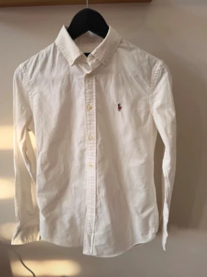 Vit skjorta från Ralph Lauren S - Klassisk vit skjorta i storlek S från Ralph Lauren med broderad logga på bröstet. Slim fit modell. Tillverkad i mjuk bomull för en clean och tidlös look.