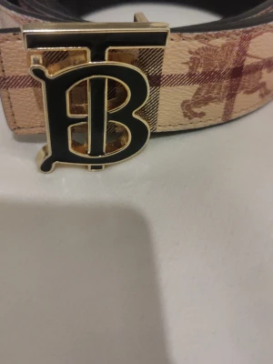 Burberry vändbart bälte med monogram - Stilrent vändbart bälte från Burberry med klassiskt rutigt mönster på ena sidan och svart på andra. Snyggt guldfärgat spänne med Burberrys monogram. Perfekt accessoar för att lyfta din outfit.