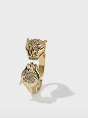 Guldfärgad ring med panterhuvuden ifrån iokiu - Unik guldfärgad ring med två detaljerade panterhuvuden i ändarna. Huvudena är dekorerade med små gnistrande stenar och har gröna ögon. Ringen har en öppen design och är tillverkad i metall med glittrande inslag för en cool och djurisk look.