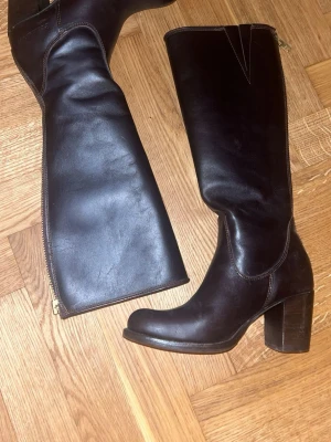 Höga mörkbruna boots  - Säljer ett par höga boots i mörkbrunt läder med markerad blockklack och rundad tå. Skorna har dragkedja längs hela skaftet och stilrena sömmar som ger en klassisk och trendig look. Nästan oanvända och från primeboots 