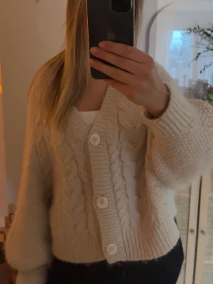 Beige kabelstickad kofta med stora knappar - Mysig beige kofta med kabelstickat mönster och tre stora vita knappar framtill. Koftan har ballongärmar och bred ribbad mudd vid ärmslut och nederkant. Knappt använd 🥰