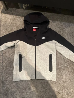 Nike Tech Fleece hoodie svart/grå - Nike Tech Fleece hoodie i svart och ljusgrått med hel dragkedja och två fickor med svarta dragkedjor. Klassisk Nike-logga på bröstet och stor huva. Perfekt för en sportig och avslappnad stil.