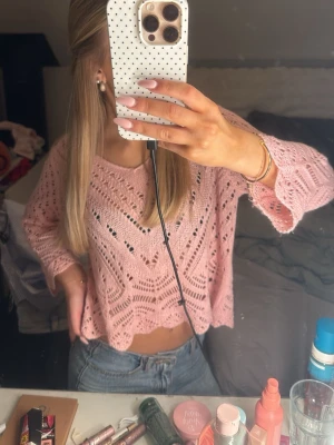 Rosa stickad cropped tröja - Söt rosa stickad tröja med hålmönster och avslappnad, croppad passform. Tröjan har långa ärmar och bred halsringning, perfekt att styla med jeans. Materialet ser ut att vara mjukt och luftigt, vilket ger en trendig och bekväm look.