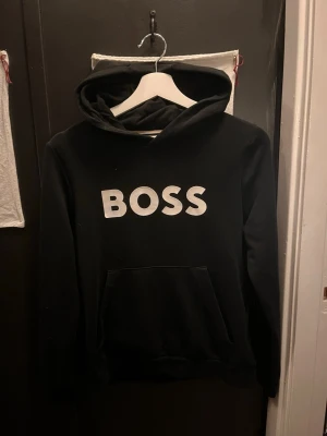 Boss Hoodie - Svart hoodie från BOSS med stor vit logga på bröstet. Klassisk modell med huva och känguruficka framtill. Perfekt för en avslappnad och stilren look. Mjuk bomullskänsla och ribbade muddar vid ärmslut och nederkant. Storlek 160cm men passar mig som är 169! Nypris runt 1000kr