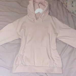 Ljusrosa Gina tricot hoodie med huva - En basic ljusrosa hoodie med stor känguruficka framtill och justerbar huva med dragsko. Hoodien har ribbade muddar vid ärmslut och nederkant. Perfekt för en chill och avslappnad stil. Materialet känns mjukt och bekvämt, troligen bomull eller bomullsblandning.
