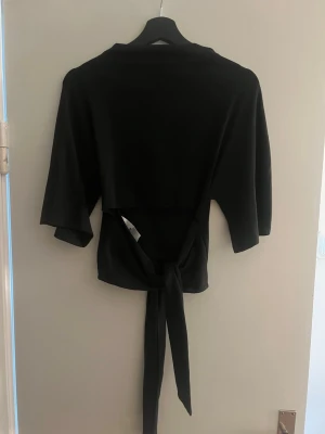 Svart topp med knyt i ryggen - Svart topp med trekvartsärm och snygg knytning i ryggen. Modellen är croppad och har en enkel, stilren look som passar till många outfits. Tillverkad i mjukt material som känns skönt mot huden.