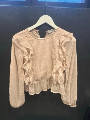 Ljusrosa volangblus med prickar från NA-KD - Supersöt ljusrosa blus från NA-KD med små vita prickar, volangdetaljer längs framsidan och vid midjan. Blusen har långärmade ärmar med resår vid ärmslut och en lätt transparent känsla. Perfekt att styla med jeans eller kjol för en trendig look.