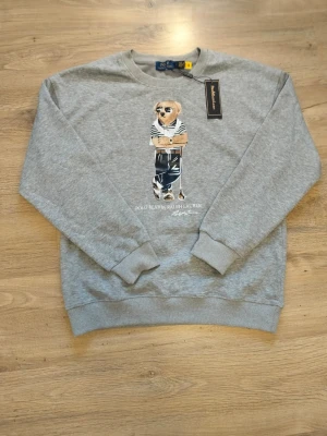 Grå sweatshirt Polo Bear Ralph Lauren - Grå sweatshirt från Ralph Lauren med ikoniskt Polo Bear-tryck framtill. Tröjan har rund halsringning, ribbade muddar och avslut. Perfekt för en avslappnad och trendig look. Det står att den är Large men den har storleken Small