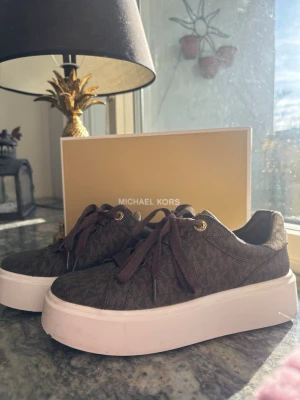 Bruna Michael Kors sneakers - Snygga bruna sneakers från Michael Kors med monogrammönster och vita platåsulor. Skorna har bruna skosnören, guldfärgade öljetter. Perfekta för dig som vill ha en trendig och stilren look med exklusiv känsla. Skorna är i storlek 37,5!🩷