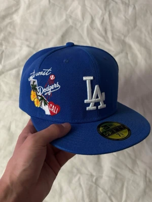 New Era keps blå - Snygg blå keps från New Era med LA Dodgers-logga framtill och broderade detaljer som 'west coast', blommor och 'CALI' på sidan. Kepsen har platt skärm och klassisk passform. Materialet är bomull och kepsen har MLB-logga baktill.