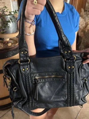 Svart handväska med flätade detaljer - Snygg svart handväska i skinnimitation med flätade detaljer på framsidan och guldiga metalldetaljer. Väskan har en dragkedjeficka framtill, två rejäla handtag och flera fack med dragkedja. Perfekt för dig som vill ha en rymlig och trendig väska.