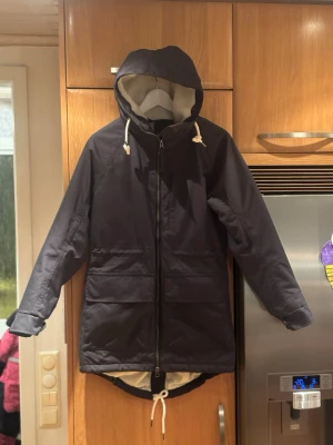 Mörkblå parkas från Tretorn med huva - Snygg mörkblå parkas från Tretorn med stor huva och dragsko i midjan. Jackan har två stora fickor framtill och är fodrad med mjuk, vit teddy på insidan för extra värme. Perfekt för kyliga dagar och har en längre modell baktill.