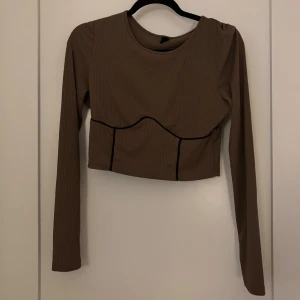 Brun croppad långärmad topp - Trendig brun croppad topp med långa ärmar och svarta kontrastsömmar. Toppen har en ribbad struktur och markerade sömmar framtill som ger en cool look. Perfekt att styla med höga jeans eller kjol.