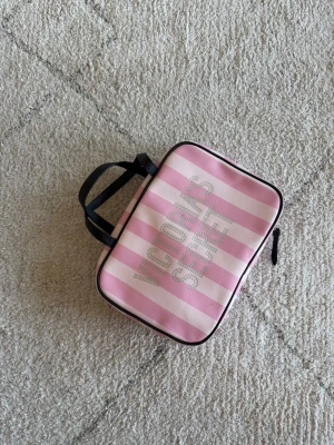 Victoria's Secret rosa randig väska - Säljer en söt väska från Victoria's Secret med rosa och vita ränder, dekorerad med glittrande stenar i loggan. Väskan har svarta handtag och är perfekt för smink eller småprylar. Materialet är tåligt tyg och formen är rektangulär.