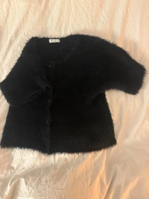 Svart fluffig kofta från Wera  - Säljer en svart, kortärmad kofta från Wera med fluffig, mjuk textur. Koftan har rund halsringning och knäppning framtill. Perfekt för dig som vill ha en mysig och trendig look med lite extra attityd.