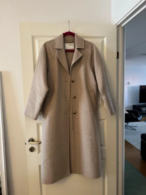 Beige ullkappa från Selected Femme - Stilren beige ullkappa från Selected Femme med klassisk krage och knäppning framtill. Kappan har raka ärmar, diskreta sidofickor och en rak, elegant passform. Perfekt för dig som vill ha en tidlös och snygg look under kyliga dagar.