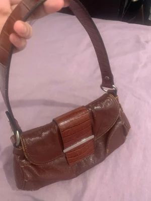 Brun axelremsväska i skinn - Snygg brun handväska i skinn med axelrem och detaljer i krokopräglat läder. Väskan har ett lock med metallspänne och en kompakt, mjuk form. Perfekt för dig som gillar vintage vibes och klassisk stil.