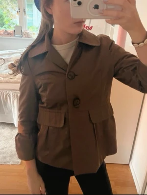 Kort brun trenchcoat med stora knappar - Säljer en kort brun trenchcoat med stora knappar framtill och bred krage. Jackan har två stora fickor med lock och är tillverkad i ett kraftigt tyg som ger en klassisk look. Perfekt för dig som gillar stilrena och tidlösa plagg.
