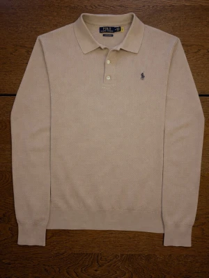 Beige långärmad pikétröja Polo Ralph Lauren - Exclusive beige långärmad pikétröja från Polo Ralph Lauren med krage, knappar och broderad logga på bröstet. Tröjan är i bomull och har en normal passform, perfekt för en clean och stilren look.