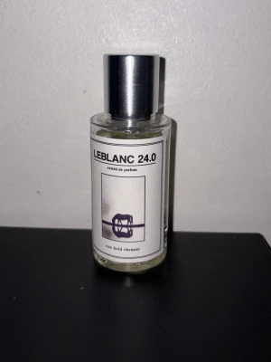 LEBLANC 24.0 parfymflaska - Stilren parfymflaska från One Bold Chemist med namnet LEBLANC 24.0. Flaskan är genomskinlig med en silvrig metallkork och har en minimalistisk vit etikett med svart text och grafisk design. Perfekt för dig som gillar unika dofter och snygg design.