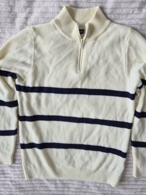 Vit stickad tröja med blå ränder - Vit ribbstickad tröja från LC Waikiki med mörkblå horisontella ränder. Tröjan har halv dragkedja framtill och hög krage för extra stilpoäng. Perfekt för dig som gillar klassisk men ändå trendig look.