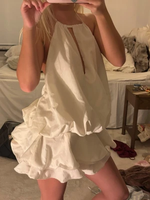 Vit volangklänning med halterneck - Supersöt vit kort klänning med halterneck och volangig kjol. Klänningen har en luftig passform och ett litet sprund framtill. Perfekt för sommarens alla tillfällen och ger en fräsch och trendig look.