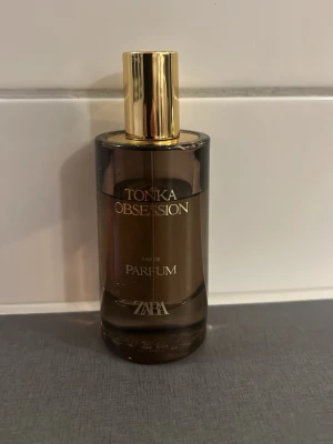 Tonka Obsession parfym från Zara - Tonka Obsession Eau de Parfum från Zara i en elegant brun glasflaska med guldfärgat lock. Flaskan har en rundad form och stilren design med guldtext på framsidan. Perfekt för dig som gillar exklusiva och varma dofter.
