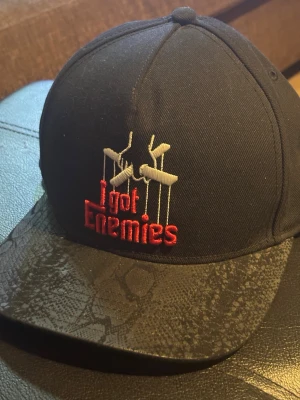 Svart snapback från Cayler & Sons - Snygg svart snapback-keps från Cayler & Sons med broderad text 'I got Enemies' i rött och vitt framtill. Skärmen har ett unikt ormmönster som ger extra attityd. Perfekt för dig som vill sticka ut med streetwear-vibe.