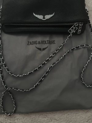 Svart axelväska från Zadig & Voltaire - Snygg svart axelväska i läder från Zadig & Voltaire med silverfärgad kedjerem och dragkedjor. Väskan har en ikonisk vingdetalj i silver framtill och en praktisk ytterficka med dragkedja. Perfekt för att lyfta din outfit.