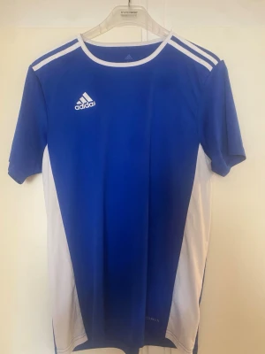 Blå Adidas sporttröja med vita detaljer - Snygg blå sporttröja från Adidas med vita paneler på sidorna och klassiska vita ränder på axlarna. Tröjan har rund halsringning och korta ärmar, perfekt för träning eller fotboll. Tillverkad i ett lätt och ventilerande material.