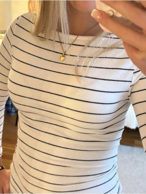 Vit långärmad randig topp - Snygg vit långärmad/trekvartsärmad topp med tunna svarta ränder. Figurnära passform och rund halsringning. Perfekt att styla med jeans eller kjol för en clean och fräsch look.