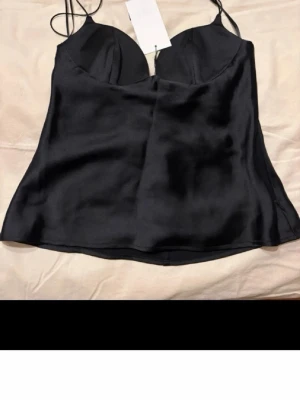 Svart satinlinnetopp med tunna band - Snygg svart linnetopp i satin med tunna axelband och lätt glansig finish. Toppen har en croppad passform och formade kupor för extra stöd. Perfekt att styla med jeans eller kjol för en trendig look.