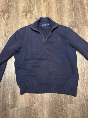 Mörkblå halvzip-tröja från Polo Ralph Lauren - Mörkblå stickad tröja med halv dragkedja och hög krage från Polo Ralph Lauren. Tröjan har ribbade muddar vid ärmslut och nederkant samt en liten grön logga broderad på bröstet. Perfekt för lager-på-lager-stil.