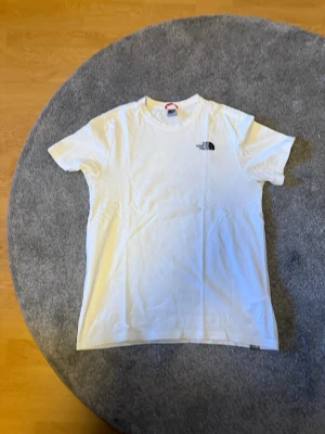 Vit t-shirt från The North Face - Snygg vit t-shirt från The North Face med liten logga på bröstet och stort grafiskt tryck på ryggen med naturmotiv. Klassisk passform och rund halsringning. Perfekt till jeans eller shorts för en avslappnad look.