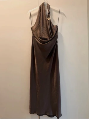Brun satin halterneck klänning - helt ny. - Snygg brun halterneck klänning. Klänningen har en dekorativ metallring vid axeln och är tillverkad i ett glansigt, mjukt material som ger ett elegant fall. Storlek XL. Helt ny klänning. 