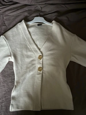 Beige kofta med guldknappar - Jättefin tröja från Gina Tricot, figursydd i midjan med två snygga knappar framtill. Säljer då den inte kommer till användning🤩