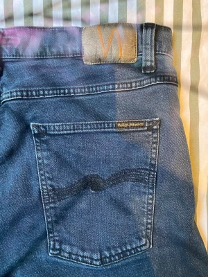  Nudie Jeans Grim Tim 36/36 Black Skies - Snygga mörkblå jeans från Nudie Jeans med klassisk femficksmodell och raka ben. Jeansen har Nudie Jeans ikoniska vågmönster på bakfickorna och en läderpatch med logga i midjan. Tillverkade i slitstark denim, perfekta för en avslappnad stil.