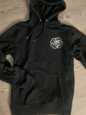 Svart hoodie med ros - Svart hoodie med stor huva och vitt tryck av en ros. Hoodien har magficka och ribbade muddar. Perfekt för dig som gillar streetwear och grafiska tryck. Aldrig använd, från trycklagret 🌹✨