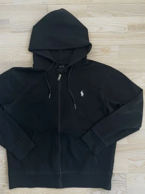 Svart hoodie från Polo Ralph Lauren - Snygg svart hoodie med dragkedja från Polo Ralph Lauren. Klassisk design med huva och dragsnören, två fickor framtill och den ikoniska vita logotypen broderad på bröstet. Perfekt för en avslappnad och stilren look.