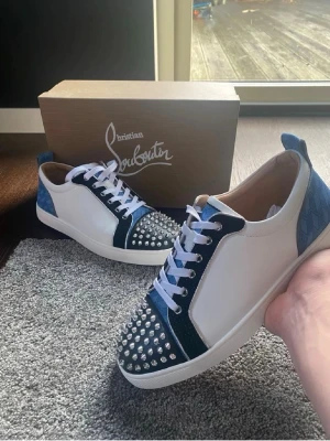 Christian Louboutin sneakers med nitar - Unika sneakers från Christian Louboutin i vitt, svart och blått. Skorna har vita snören, blå detaljer i mocka och skinn samt silverfärgade nitar på tån. Snygg kontrast mellan material och färger, med klassisk Louboutin-stil och coola detaljer.