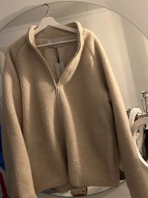 Beige teddyjacka från Norvie - Mysig teddyjacka i beige från Norvie med dragkedja framtill och hög krage. Jackan har en fluffig, fårullsliknande yta och är perfekt för att hålla värmen. Enkel och stilren design som passar till det mesta. Materialet är mjukt och ger en avslappnad vibe.