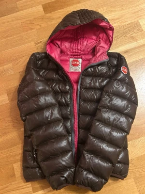 Brun pufferjacka från Colmar - Snygg brun pufferjacka från Colmar med rosa foder och huva. Jackan har quiltad design, dragkedja framtill och Colmar-logga på ärmen. Perfekt för lite kallare vår dagar eller sommar dagar. Och riktigt skön att bära. Materialet är glansigt och ger en modern look. Inga defekter! Pris går att diskutera.