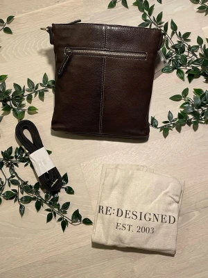 Brun läderväska crossbody äkta skinn | Re:Designed | Ny - Säljer denna tidlösa och välgjorda crossbody-väska från det danska märket Re:Designed (est. 2003). Väskan är tillverkad i äkta skinn och har guldfärgade detaljer — ett klassiskt och hållbart val. Kommer med dustbag och avtagbar axelrem. Originallappar kvar, helt oanvänd. Märke: Re:Designed Modell: MOA Small Färg: DK Brown (mörkbrun) Material: Äkta läder Skick: Helt ny, aldrig använd Ordinarie pris: 1700 kr Höjd: 25 cm Bredd: 24 cm Djup: 5 cm Axelremslängd: 140 cm (justerbar) Frakt tillkommer.