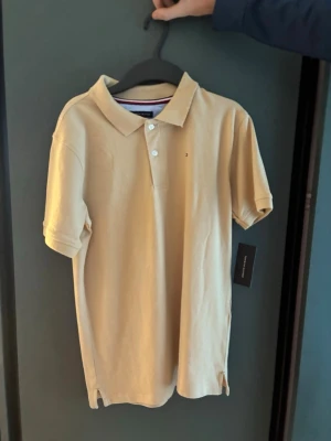 Beige pikétröja från Tommy Hilfiger - Snygg beige pikétröja från Tommy Hilfiger med klassisk krage och korta ärmar. Tröjan har knappar framtill och en liten broderad logga på bröstet. Perfekt för en stilren och avslappnad look.