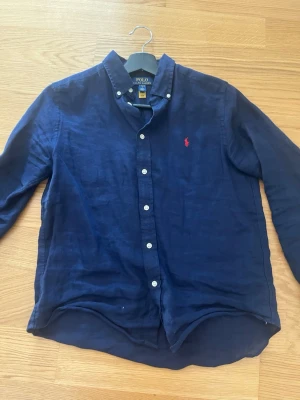 Mörkblå linneskjorta från Polo Ralph Lauren - Snygg mörkblå skjorta i linne från Polo Ralph Lauren med klassisk krage och knäppning framtill. Skjortan har långa ärmar och den ikoniska röda loggan broderad på bröstet. Perfekt för en stilren och avslappnad look.
